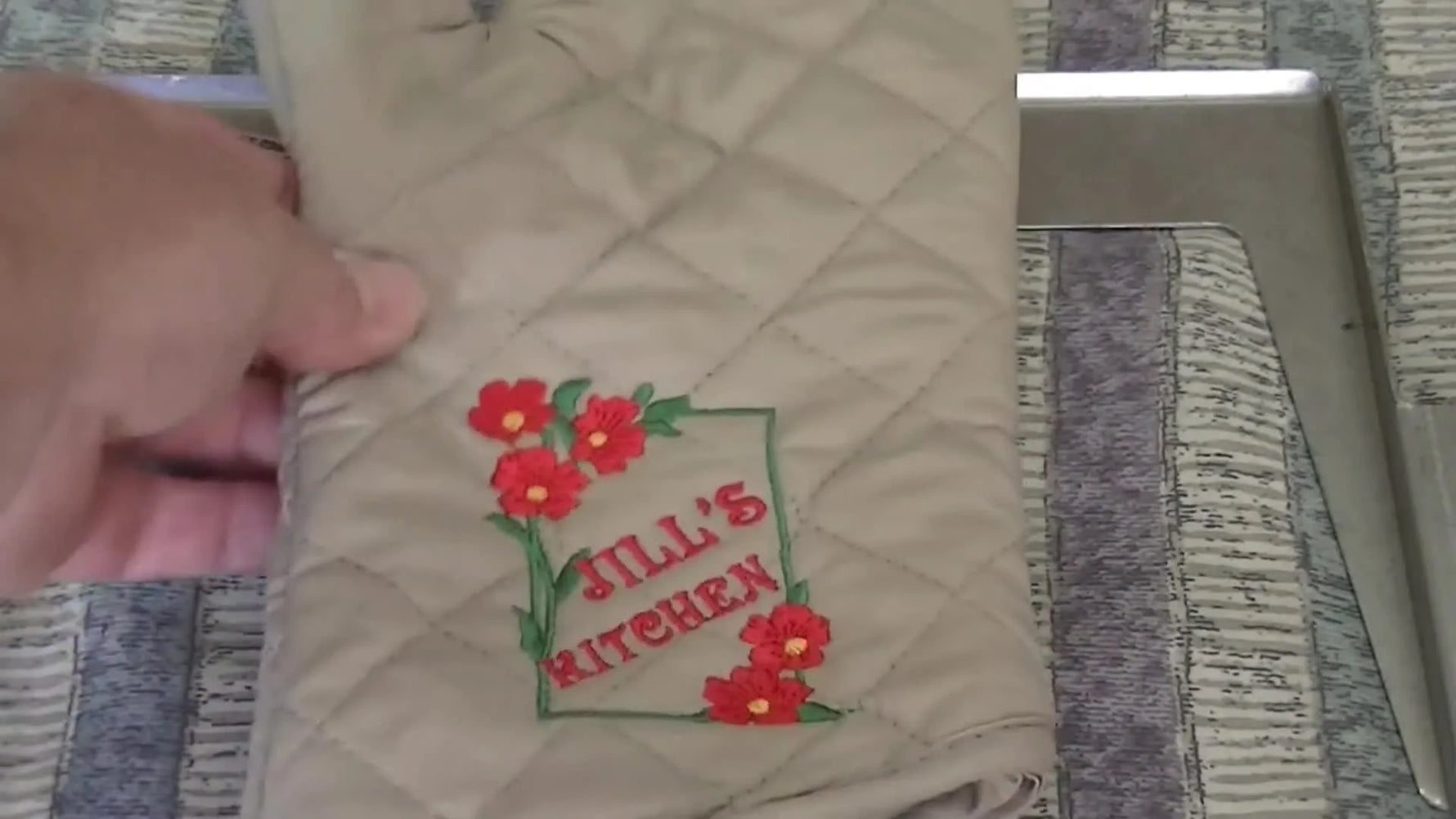 A finished quilted oven mitt is held up close to the camera, showing a floral design and the text “Jill’s Kitchen.”