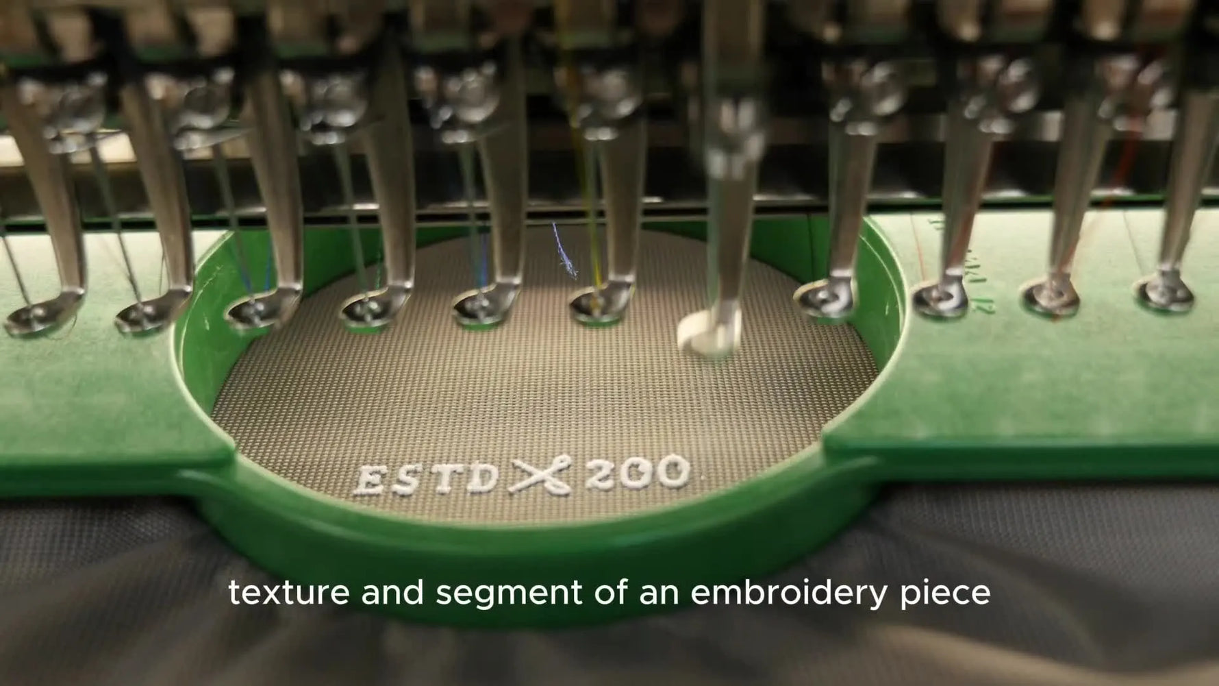 A commercial multi-needle embroidery machine stitches a logo at speed inside a green tubular hoop, showing what production-ready embroidery looks like in motion.