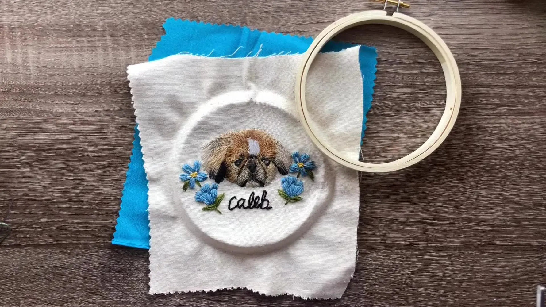 Overhead shot showing an embroidered pet portrait in an oval hoop, a piece of blue backing fabric, and an empty outer hoop ring on a wooden table.