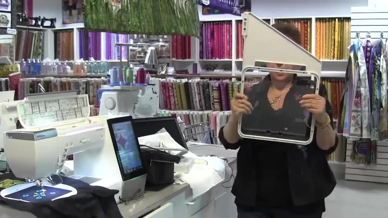 The presenter holds the Janome MC15000 quilting hoop and points out the magnets used to secure an 8x8 quilt block.