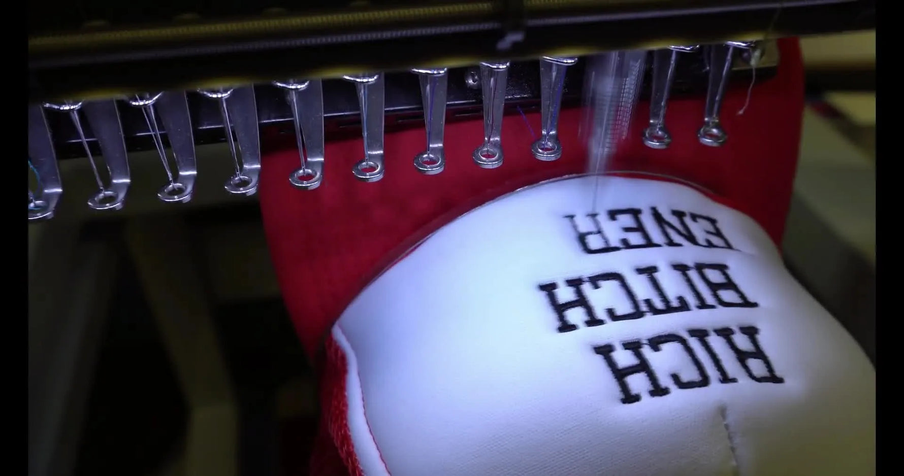 A close-up action shot of black lettering stitching at speed on a red foam trucker hat mounted on the cap driver.