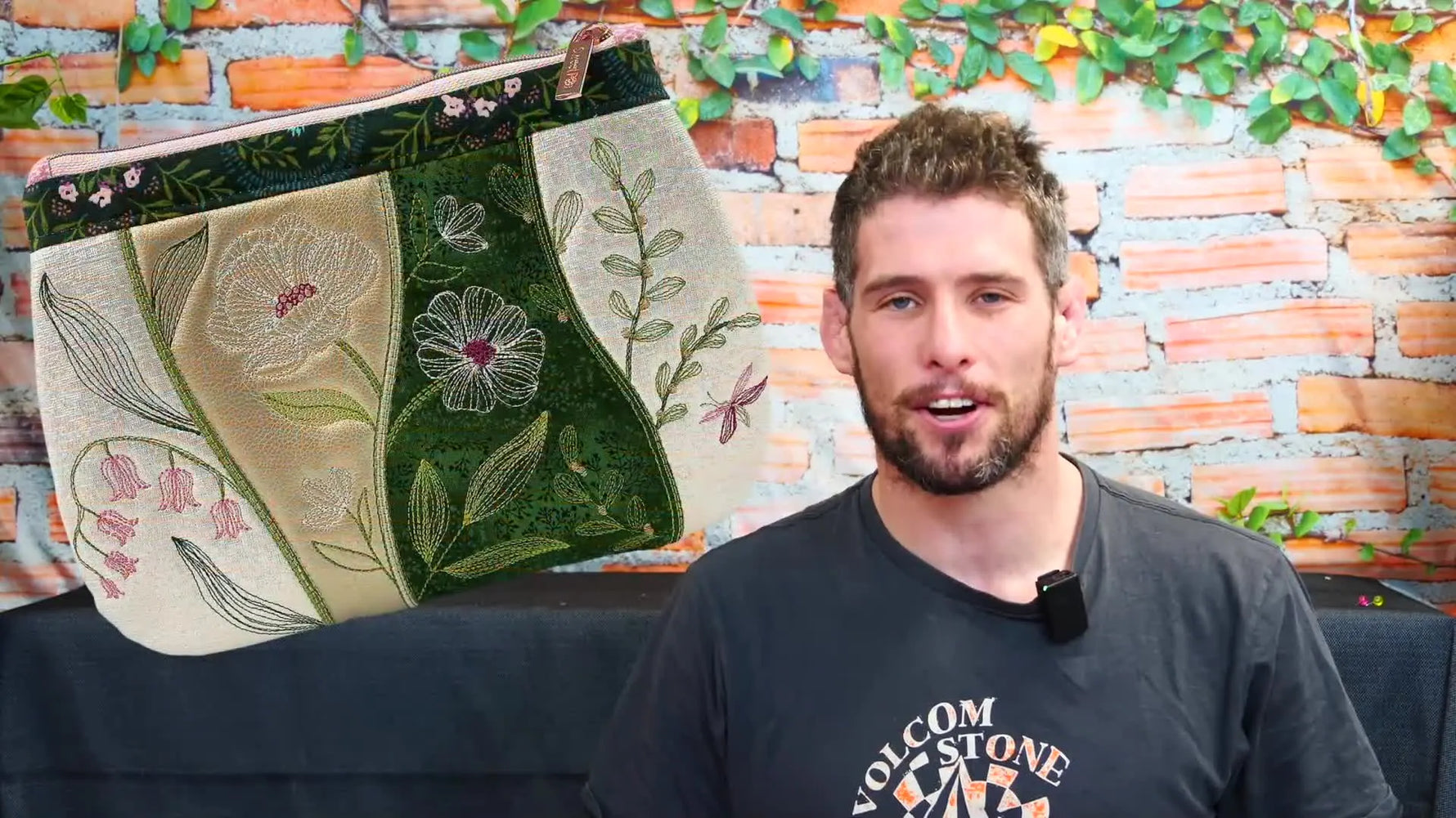 A finished floral embroidered clutch is displayed beside the presenter against a brick wall background.