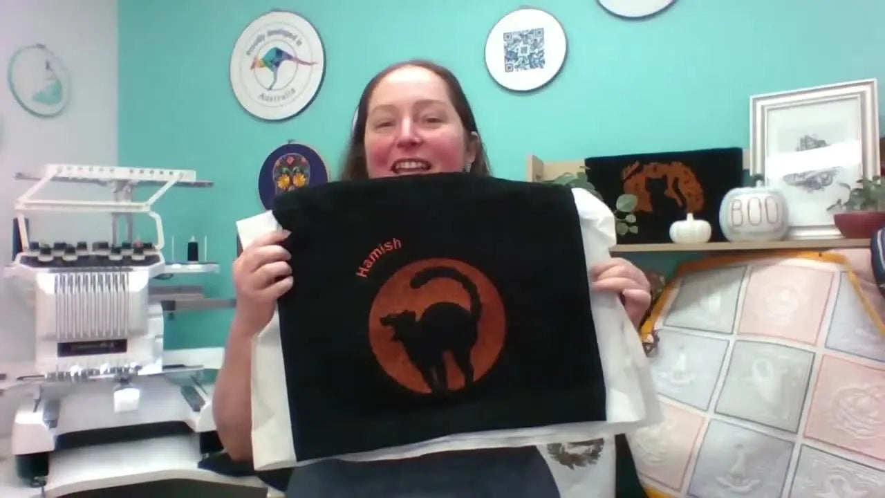 The host holds up the finished black towel, showing a Halloween design with bright lettering that stays crisp thanks to a laydown stitch base.
