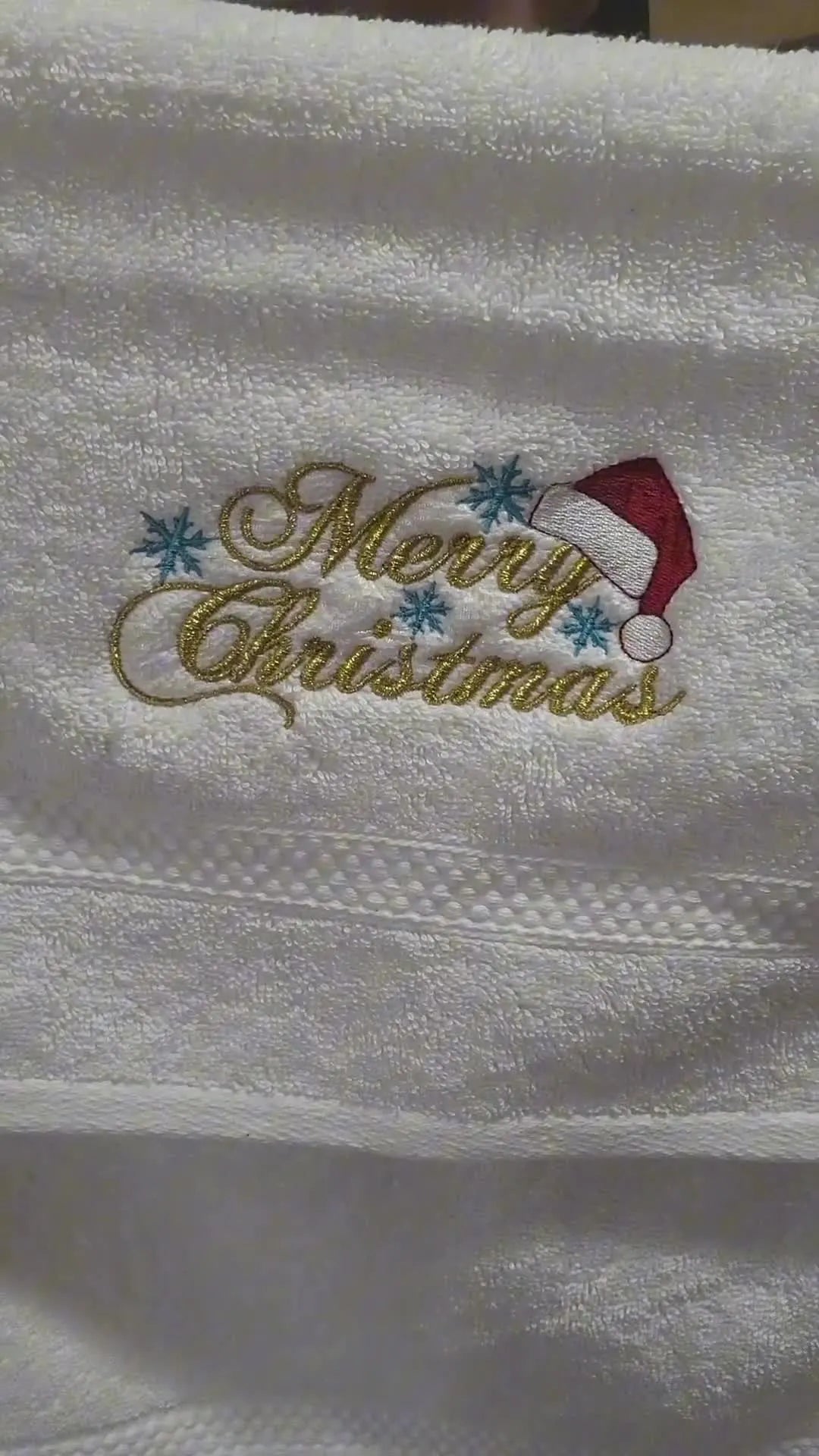 A close-up of the finished white towel featuring a clean, colorful “Merry Christmas” embroidery with crisp stitches sitting on top of the towel pile.