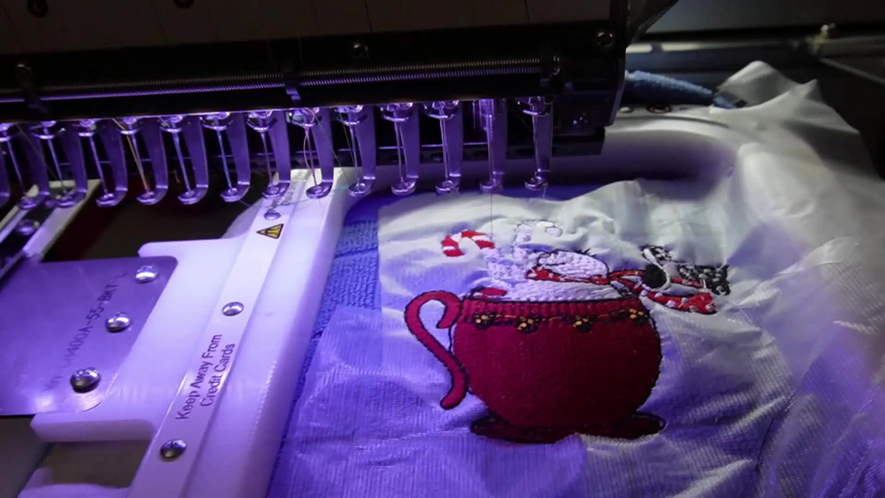 A finished “Fueled by Coffee & Christmas” snowman design sits cleanly stitched on a blue waffle weave towel while still mounted in a vertical magnetic hoop on the embroidery machine.