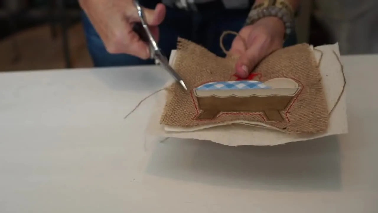 A finished rustic manger ITH ornament is held up to the camera, showing the raw-edge appliqué look and hanging loop.