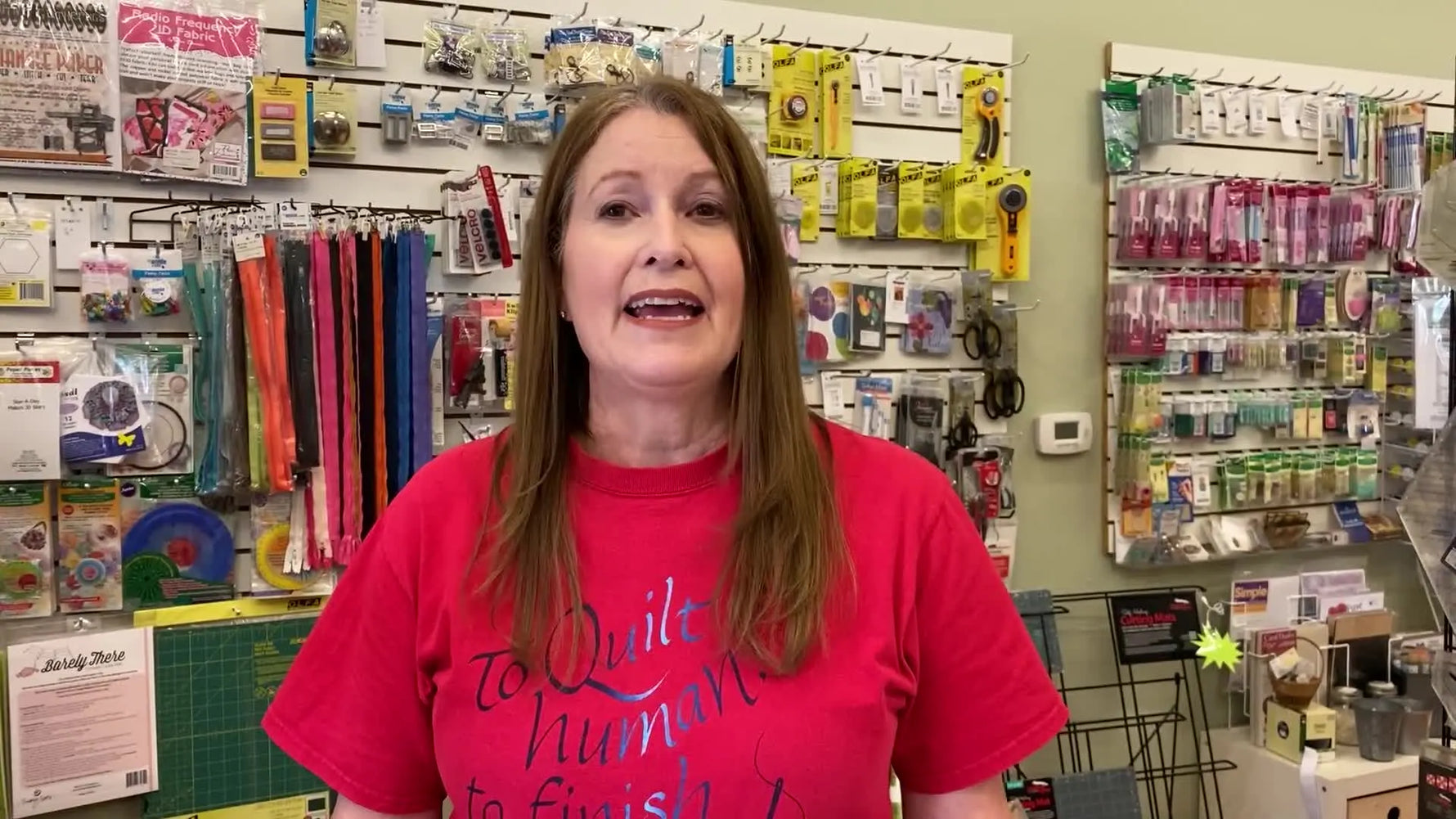 A woman in a red t-shirt stands in front of a wall of craft supplies, smiling and looking directly at the camera.