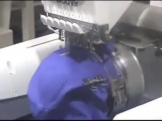 A close-up of a multi-needle embroidery machine stitching a bright logo onto a blue cap mounted in a mechanical cap frame.