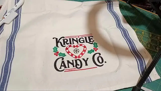 An overhead view of a finished “Kringle Candy Co.” embroidery design stitched on a striped cotton kitchen towel laid flat on a gridded cutting mat.