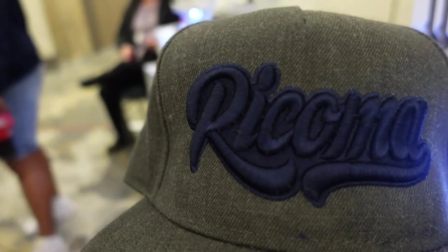 A finished gray snapback hat with a navy blue 'Ricoma' logo in 3D puff embroidery.