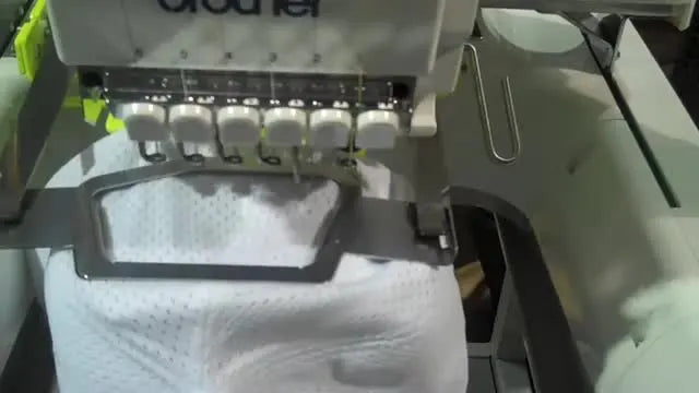 A Brother multi-needle embroidery machine stitches a phone-number design onto the side of a baseball cap held securely in a Hoop Tech window clamp.
