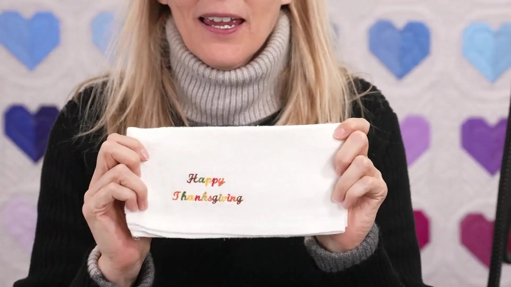 The finished double gauze napkin is held up to show crisp, multi-color “Happy Thanksgiving” lettering after stitching, tear-away, and pressing.