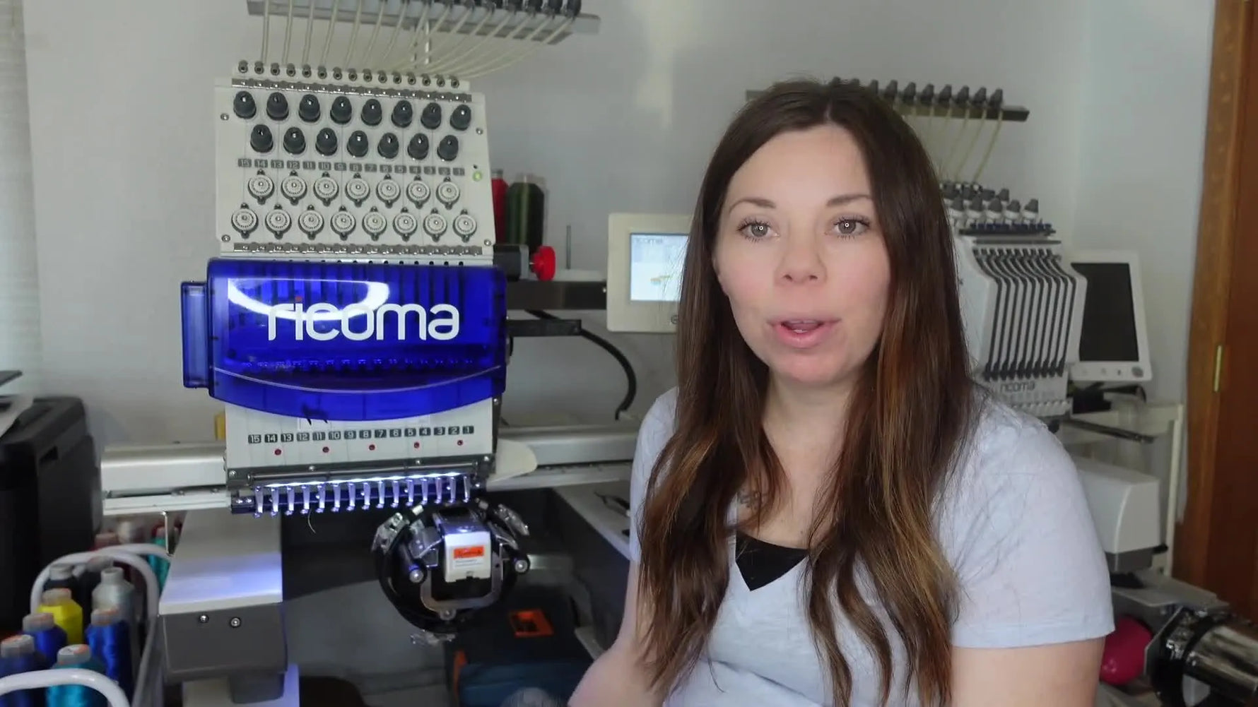 Woman holding up a grey Flexfit hat in a cap frame, smiling at the camera, with a Ricoma TC embroidery machine in the background.