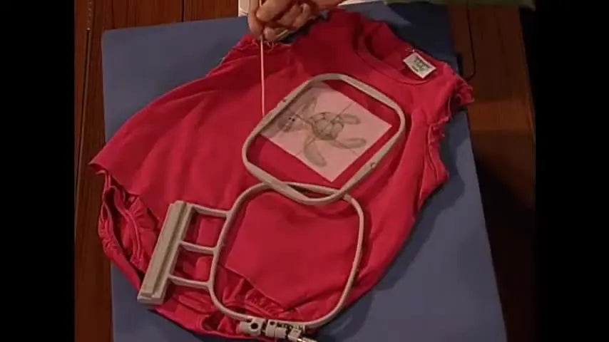 Hands place a taped inner embroidery hoop onto a red knit baby onesie using a paper template for perfect alignment.