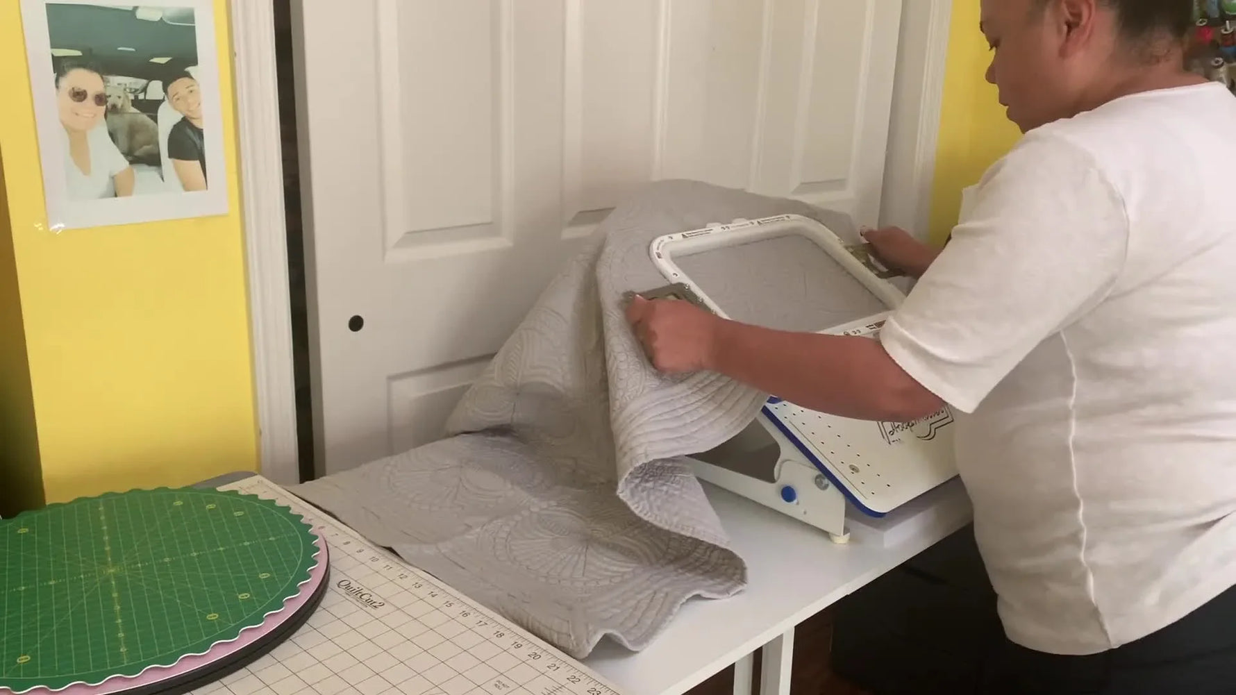 Hands snap an 8x13 magnetic hoop closed over a thick gray baby quilt blanket, creating a secure sandwich ready for precise name placement.