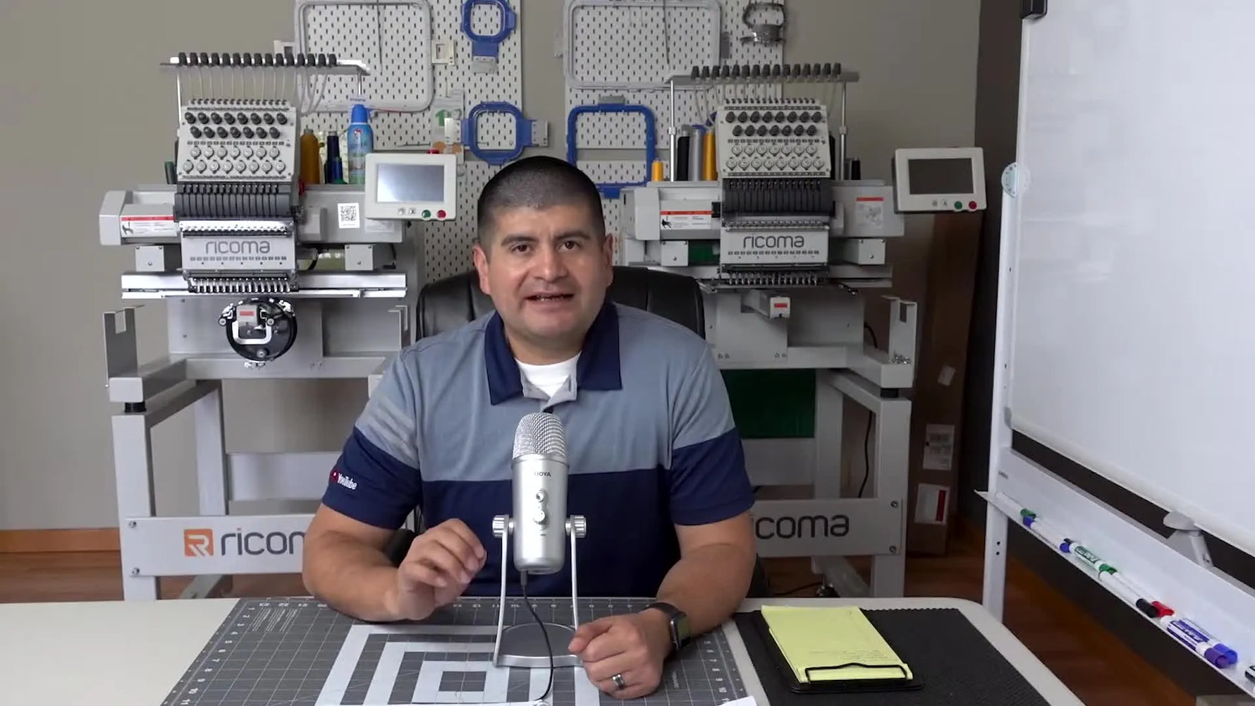 A home embroidery shop desk setup with two Ricoma multi-needle embroidery machines visible behind the speaker, signaling a production-focused workflow.