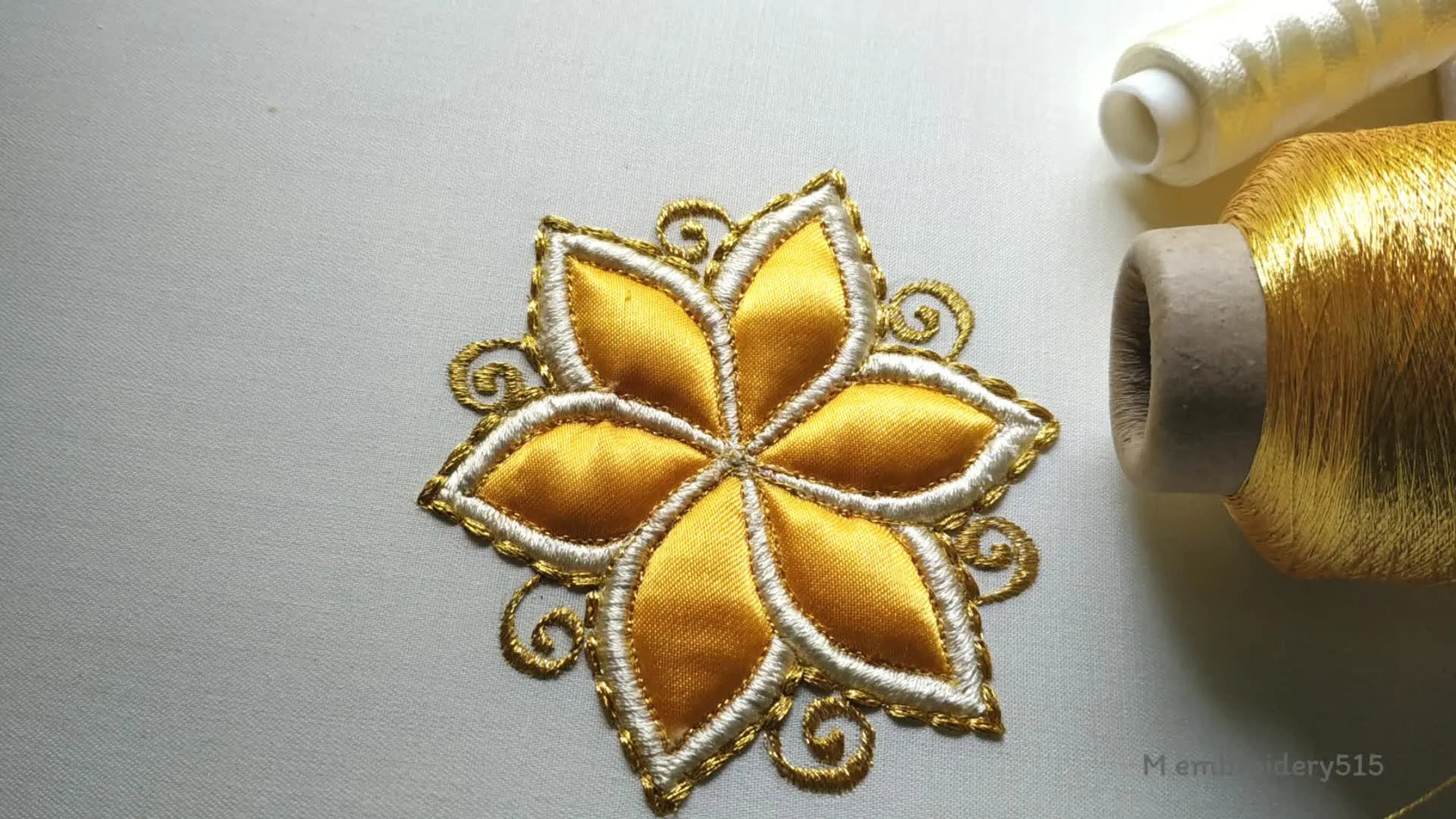 Completed golden applique flower with ornate gold thread scrollwork on white fabric, with embroidery machine and thread spools in the background.