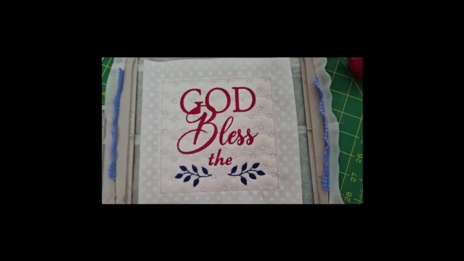 An overhead view of the finished “God Bless the” ITH quilt block still hooped, showing the quilted background and red/blue lettering.