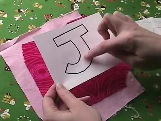 Hands holding a 'J' template over patterned fabric, with pale pink fabric underneath.