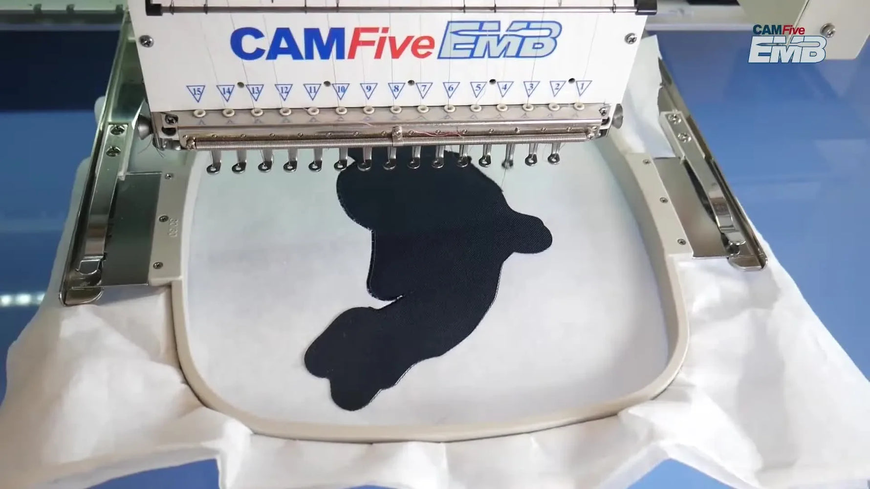 A close-up of a multi-needle embroidery machine stitching dense white details onto a dark patch base inside the hoop.