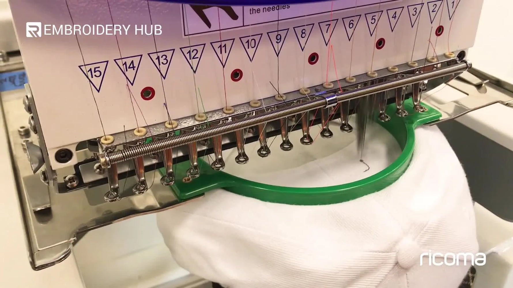 Close-up of needles stitching a logo onto the back of a white cap while it’s held in a standard green hoop.