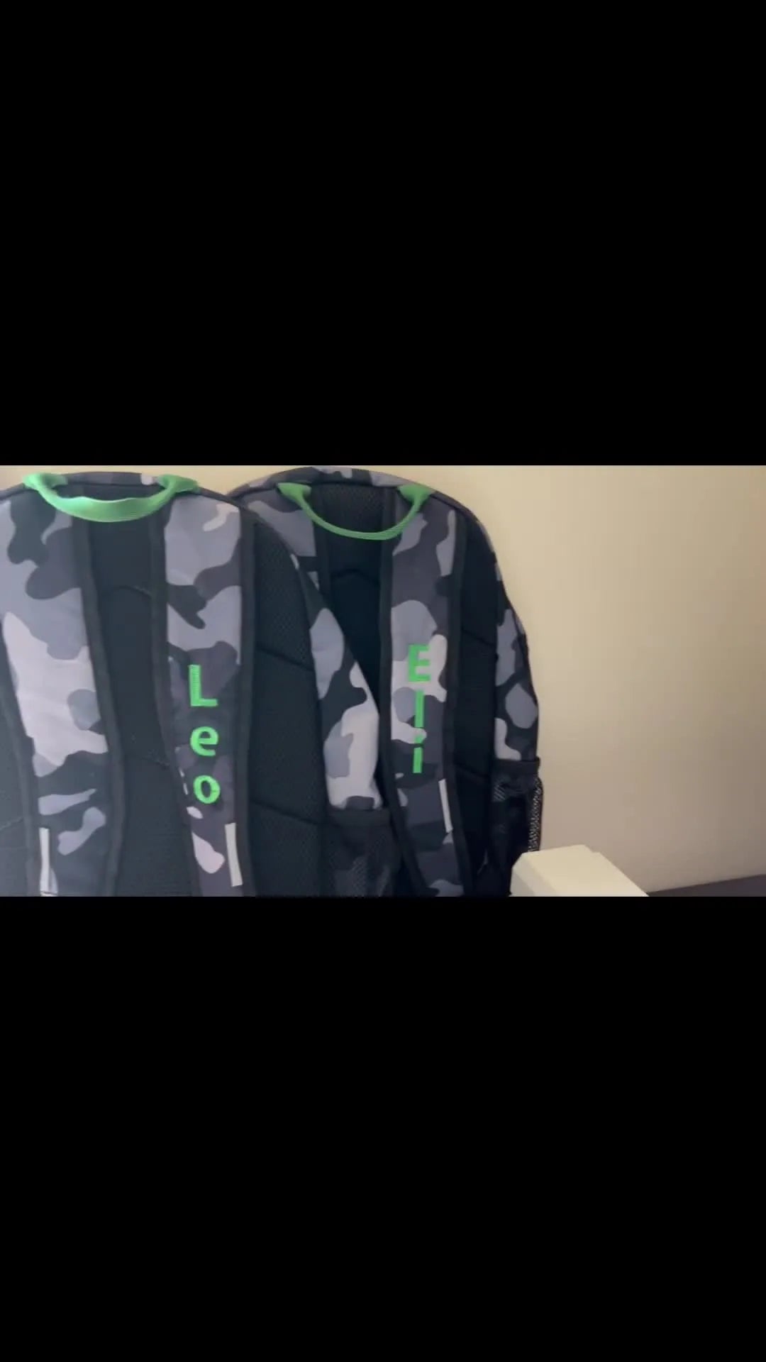 Two finished camouflage backpacks with green-trimmed straps show clean, readable names stitched in matching green thread.