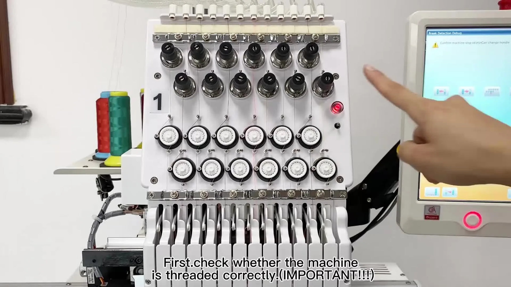 A close-up of a 12-needle embroidery machine head where the operator points at the tension area before diagnosing thread-break detection.