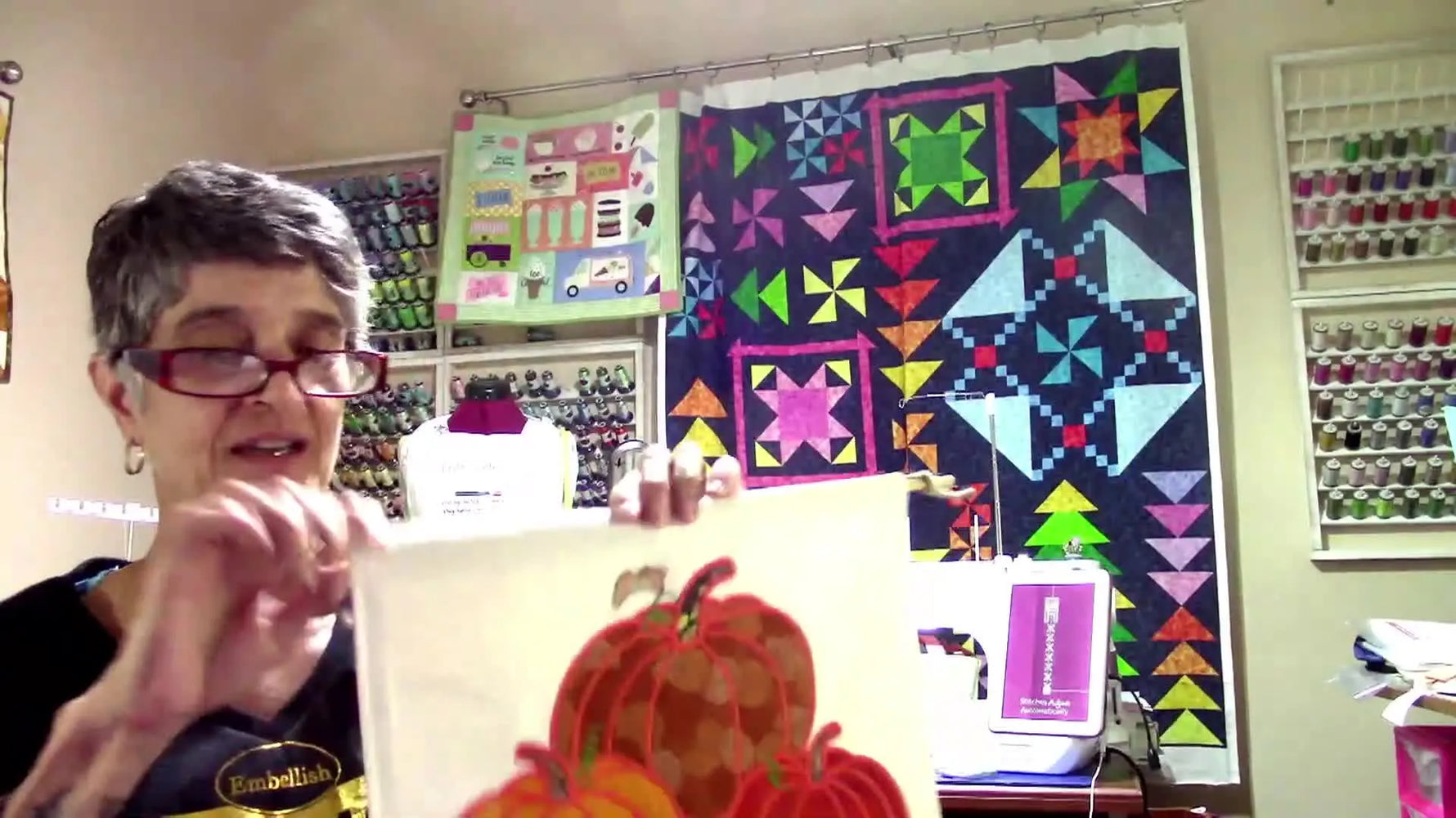 The host holds up a large pumpkin wall-hanging panel that exceeds many machines’ maximum hoop size, setting up the resizing lesson.