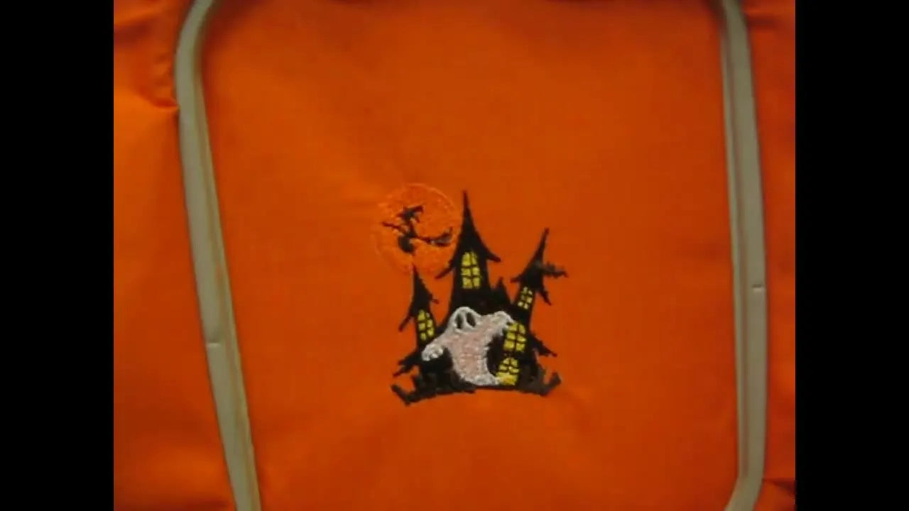 A finished haunted house embroidery design is shown completed in the hoop on orange fabric, revealing crisp color layers and clean detail.
