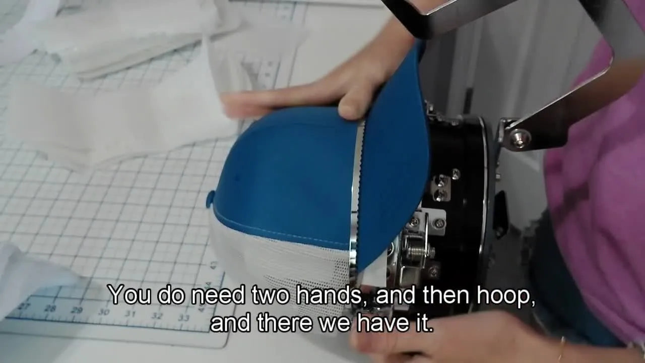 A structured snapback hat sits fully hooped and centered on the cap station, showing a flat, stable embroidery field ready for the machine.