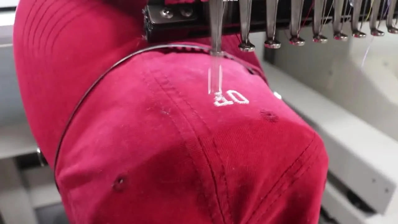 Close-up of a multi-needle embroidery machine stitching white thread onto a red dad hat during a cap-embroidery run.