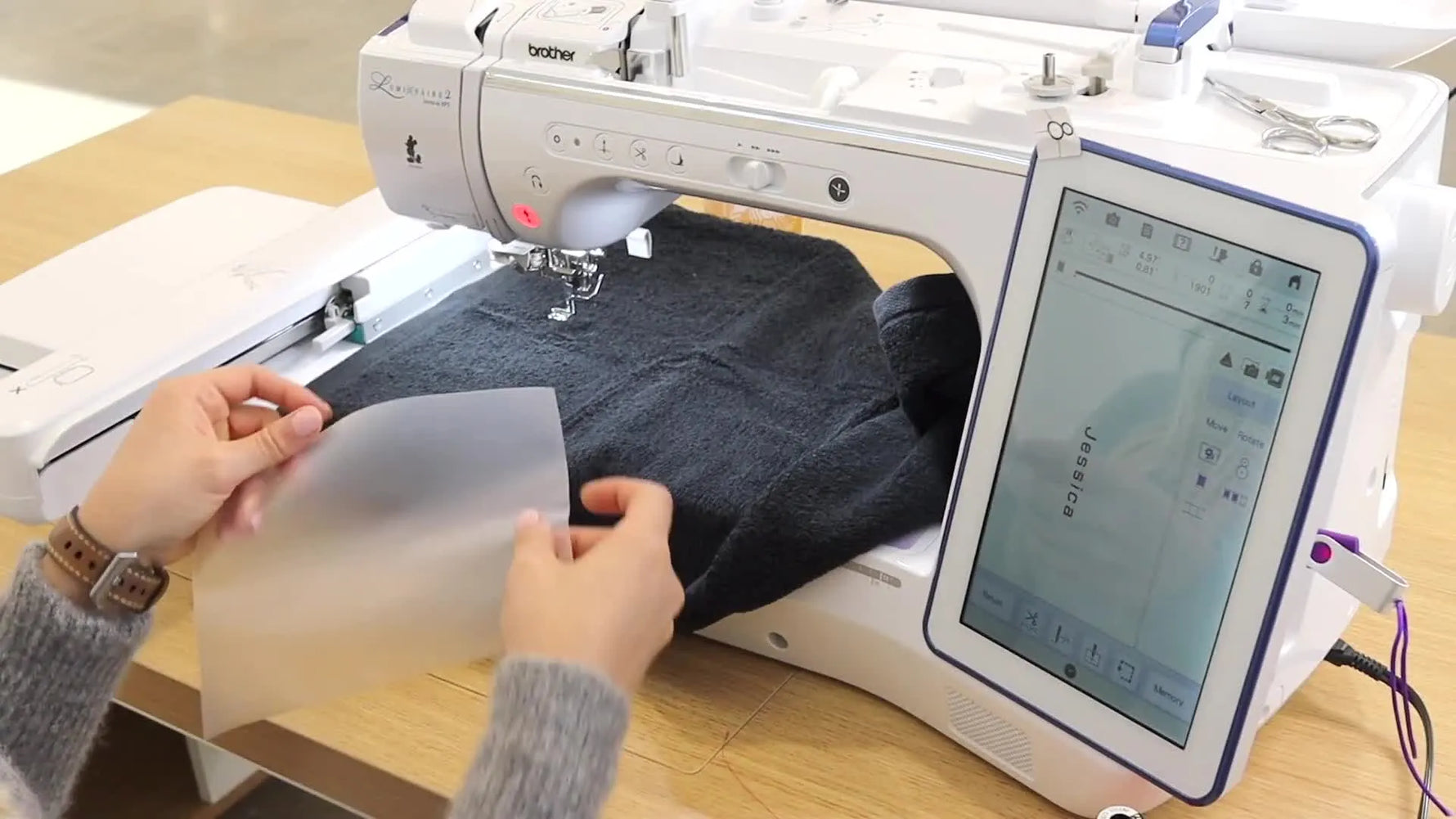 A close-up shows a clear topper being placed over a plush black towel in the hoop area right before stitching begins.