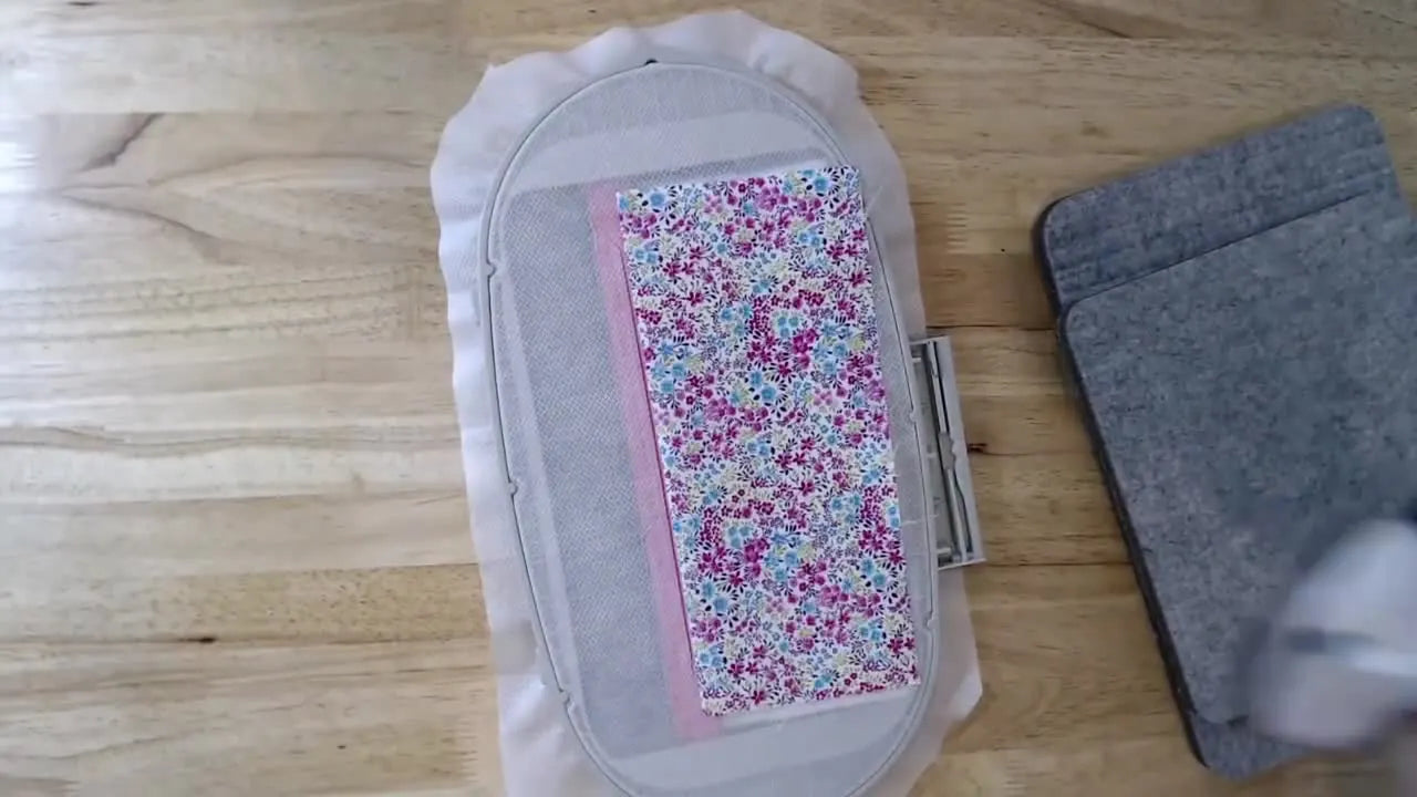 A mini iron presses a fabric fold while the embroidery hoop sits upside down on a thick non-slip press pad for stable in-the-hoop work.