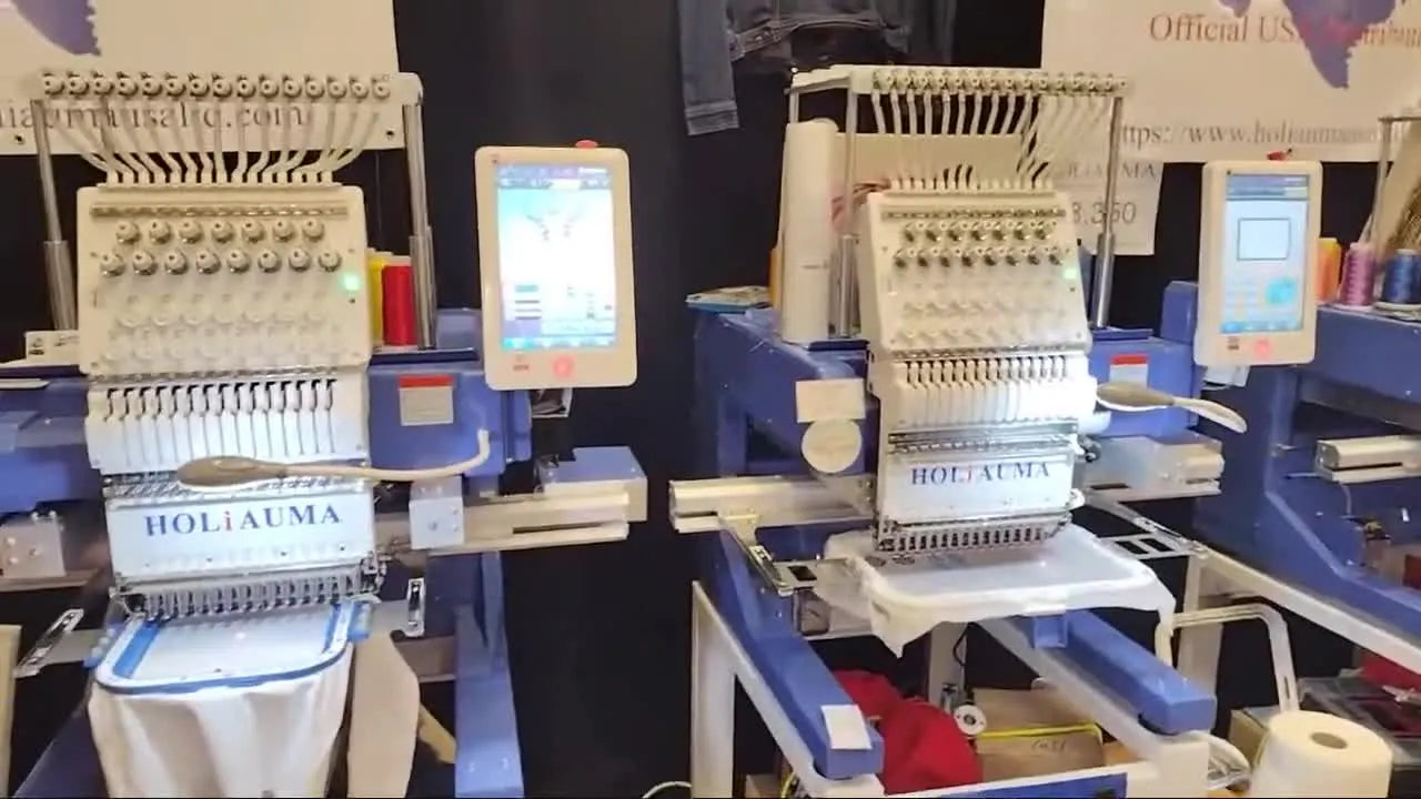 A wide booth view of a Holiauma multi-needle embroidery machine with its control screen, set up for a live stitch-out at Applique Getaway.