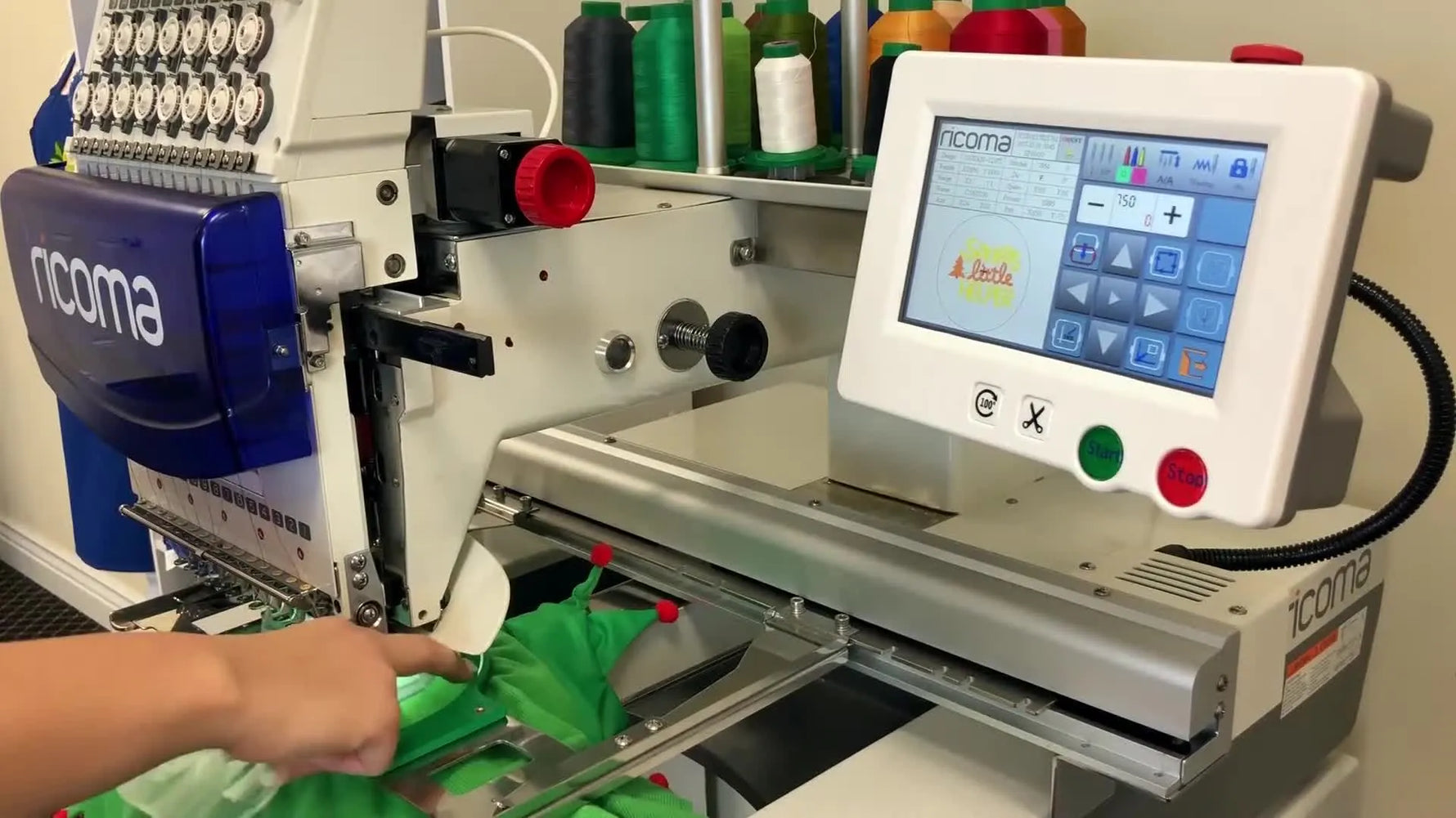 A close-up, high-angle view of a Ricoma machine stitching on a hooped green dog shirt with a clear water-soluble topping visible under the presser foot.