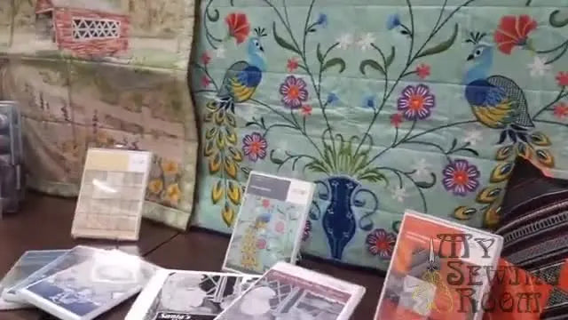 A wide display wall of finished OESD tiling scene samples, including the Peacock Tapestry, showing the scale and visual impact you can achieve with tiled embroidery blocks.