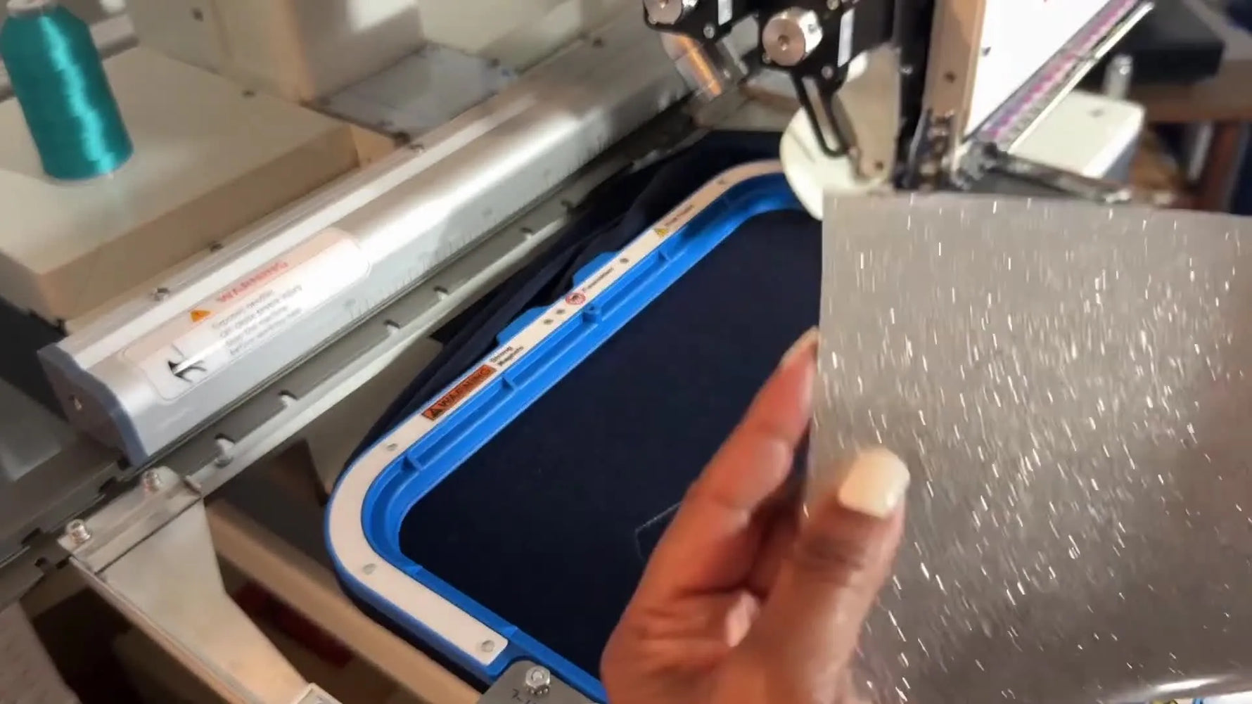 Hands peel the clear carrier sheet off glitter HTV so the vinyl can tear away cleanly after embroidery.
