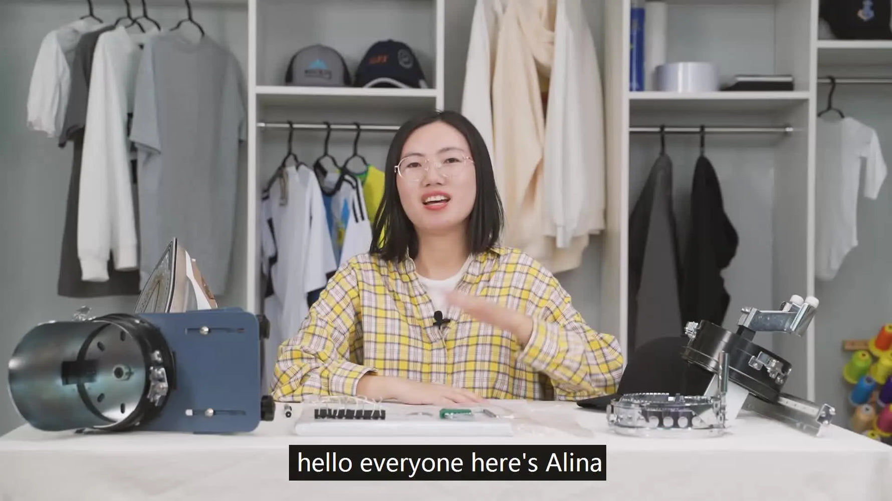 Woman in plaid shirt waves to camera, surrounded by embroidery tools and clothes racks.
