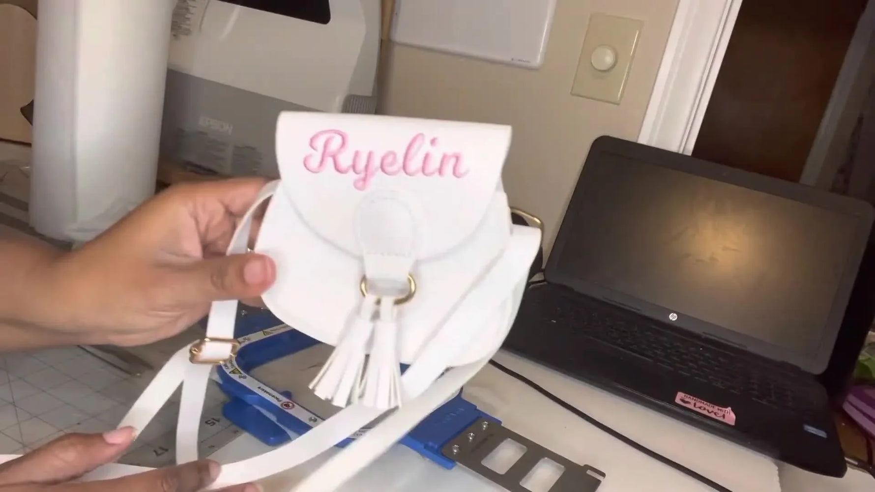 A finished white faux leather mini purse is held up to show a clean pink embroidered name centered on the flap.