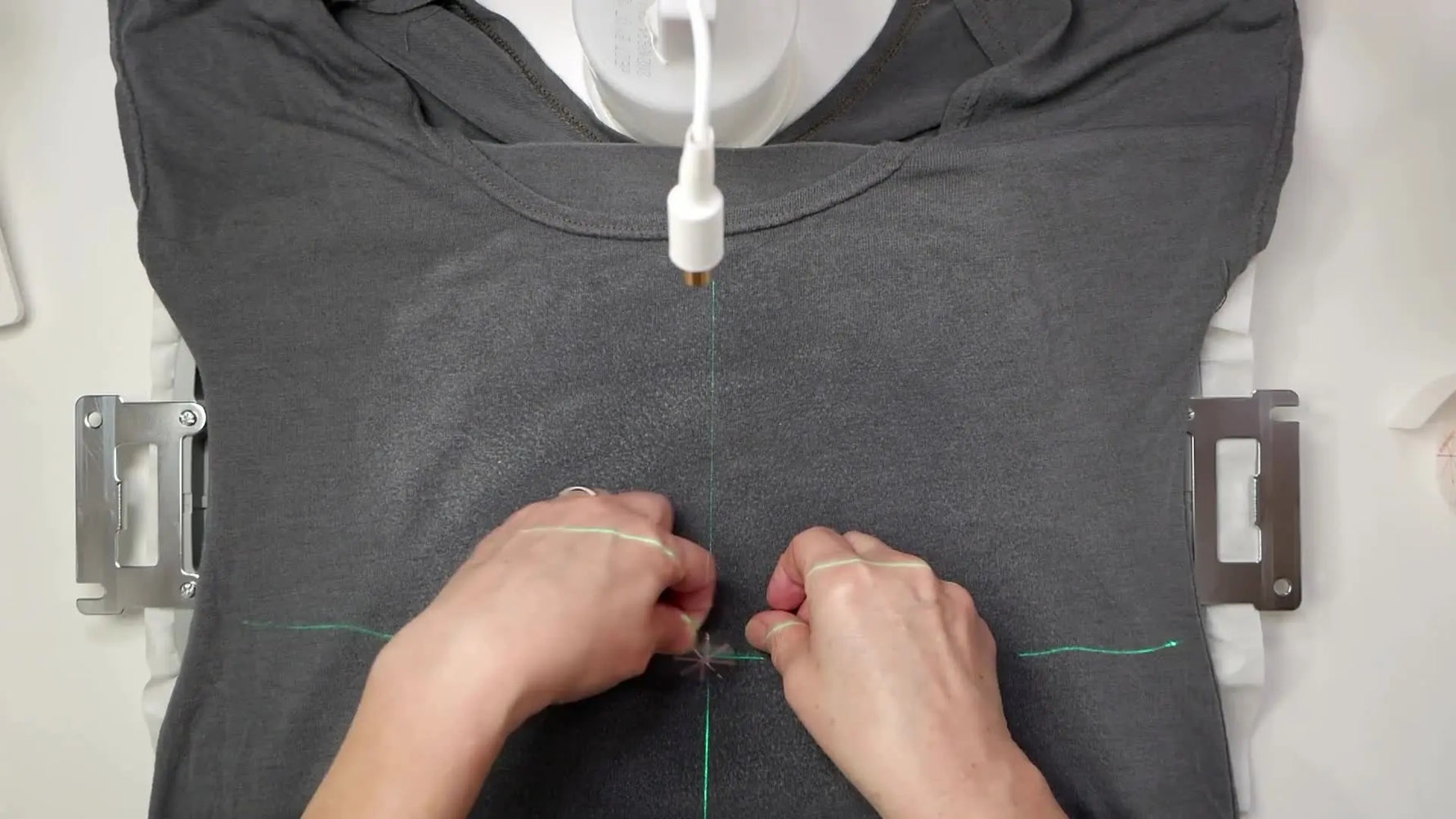 A green laser crosshair is aligned with an embroidery hoop’s grid marks while a T‑shirt is floated onto sticky stabilizer for precise design placement.