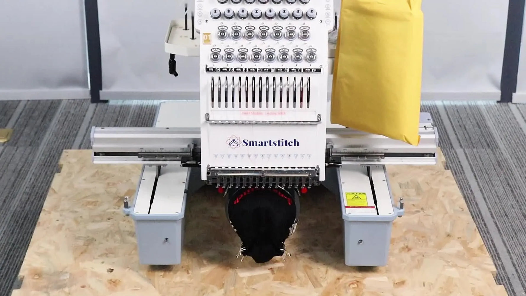 A full frontal view of the unpacked SmartStitch 15-needle embroidery machine sitting on its pallet base with the yellow cover still in place.