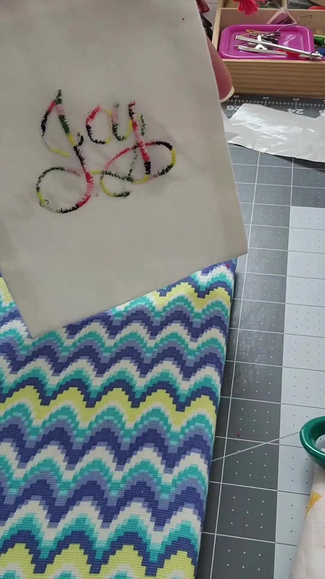 A finished canvas drawstring bag is held up to show the completed floating “Joy” iron-on embroidery patch.