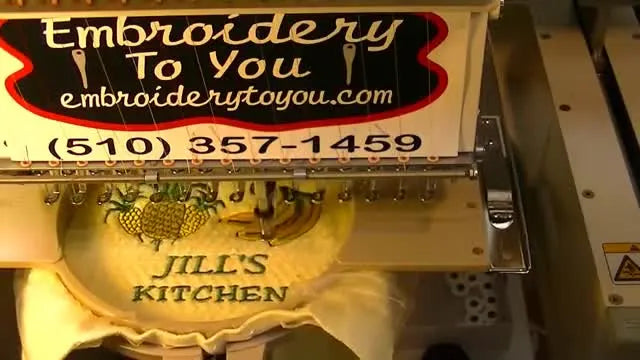 A finished waffle-weave kitchen towel sits in the hoop with crisp black text (“Jill’s Kitchen”) under pineapple-and-banana embroidery.