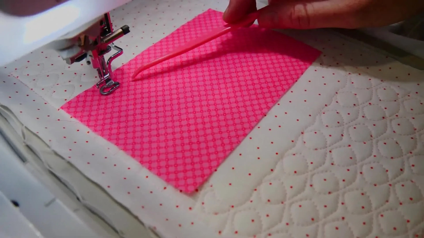 A close-up of a pink stiletto tool holding appliqué fabric flat as the needle stitches the tack-down line.