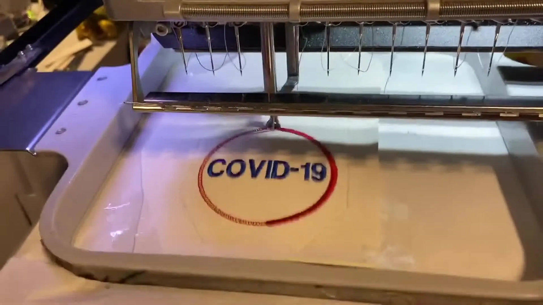 A close-up of the Melco stitching the final COVID-19 text and red circle through the layered vinyl and stabilizer.