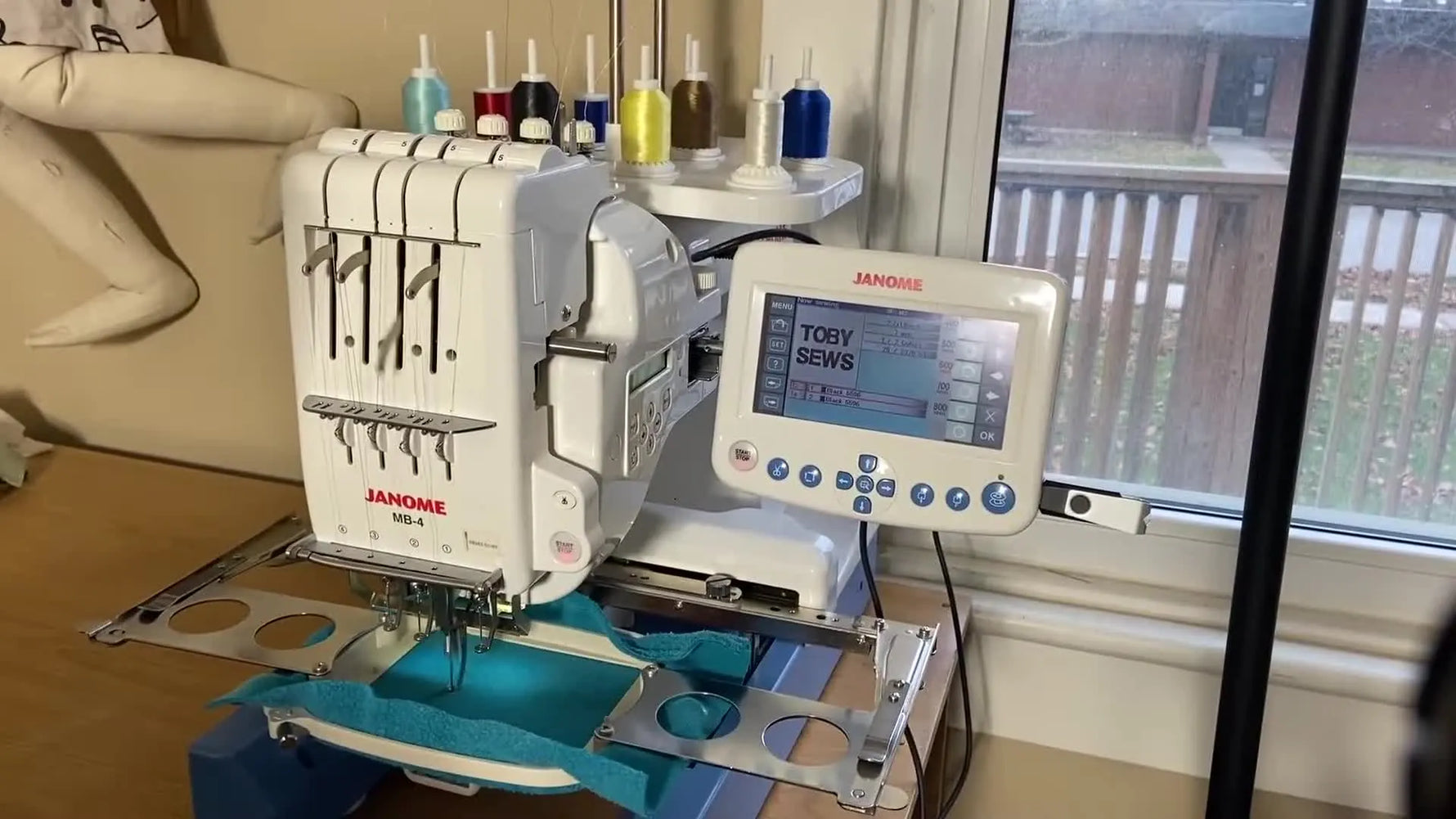 Close-up of the Janome MB-4 needle bar stitching a yellow monogram on teal sweatshirt fabric during active embroidery.