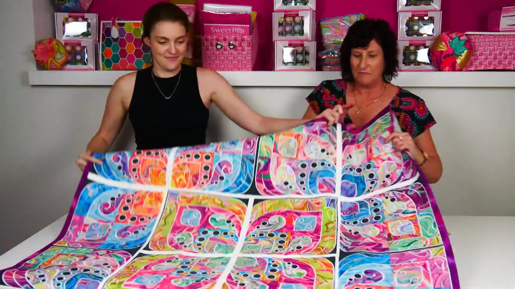 Two hosts hold up the finished Bloom Delight quilt so you can see how contrasting fat quarters and block layout choices change the final impact.