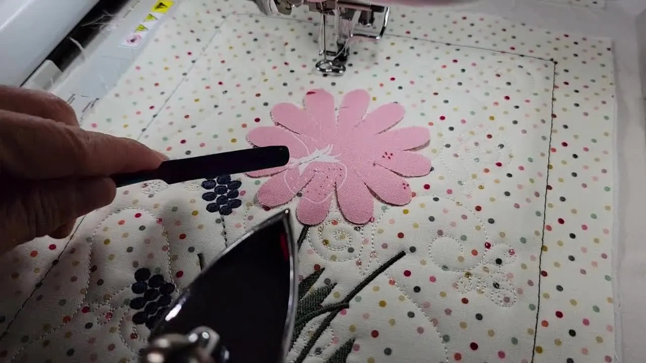 A close-up shows a small iron fusing a pink appliqué petal in the hoop while a metal spatula holds the edge safely in place.