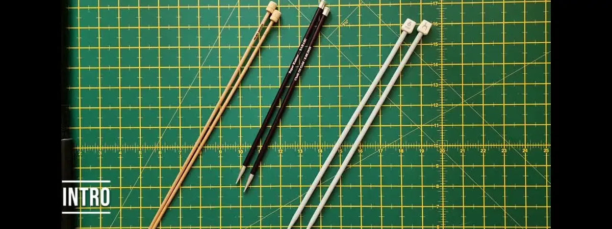 Three pairs of straight knitting needles (bamboo, metal, plastic) laid out on a green grid mat.