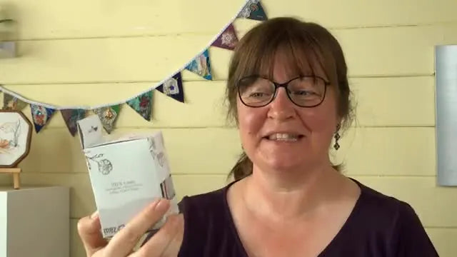 A woman with glasses and brown hair holding up a small white box of Anchor linen embroidery thread.